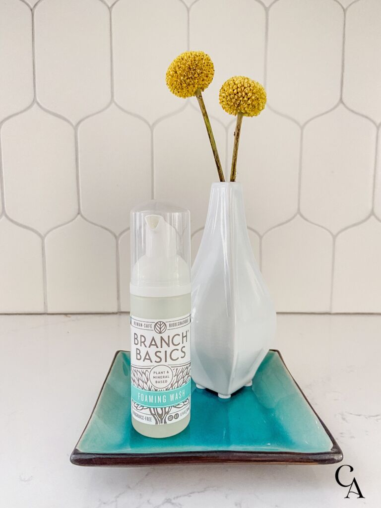 A travel sized bottle of hand soap on a turquoise plate, with a white vase of yellow flowers.