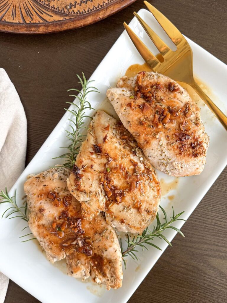 Cooked chicken with brown garlic sauce and fresh rosemary sprigs on a white plate.