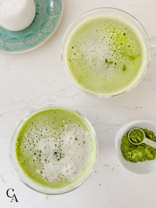 Two coconut matcha lattes on a marble countertop, next to a dish of matcha powder.