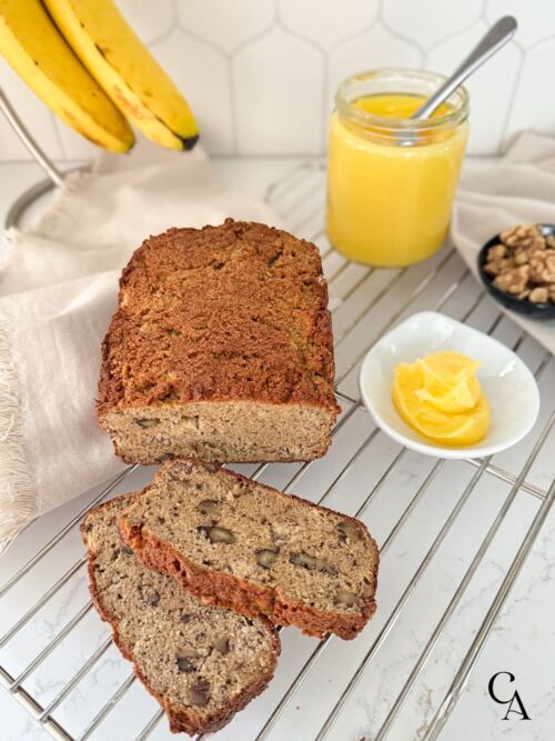 A loaf of banana bread cooling on a wire rack, next to a jar of ghee.