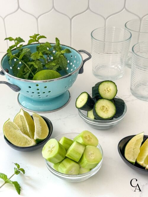 A colander of fresh mint leaves, and small bowls of sliced cucumber and lime wedges on a white counter.