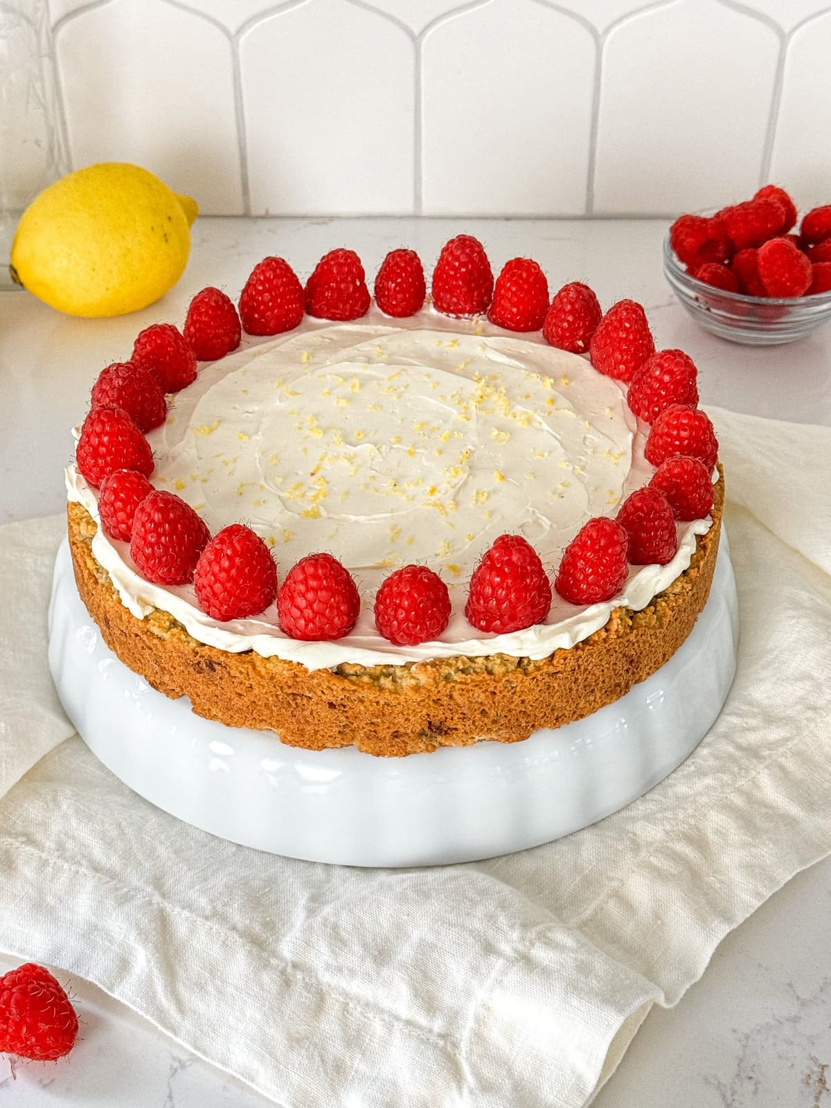 A lemon raspberry cake with cream cheese frosting, fresh berries, and lemon zest on a white dish.
