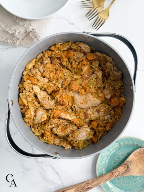 A pot of chicken vindaloo on a marble counter with a stack of plates and gold forks sitting next to it.