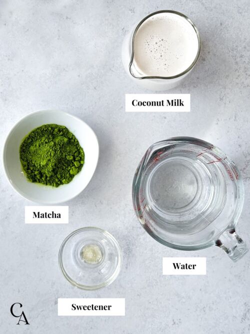 A measure cup of water, a creamer of coconut milk and a bowl of matcha powder on a grey countertop.