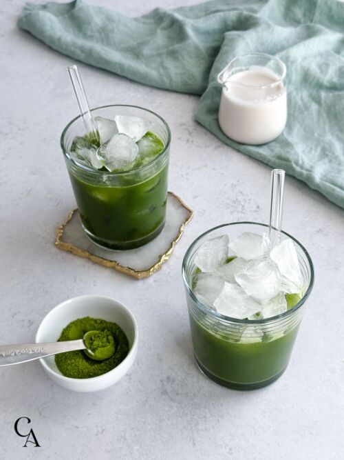 Two green drinks with ice and a small creamer of almond milk.
