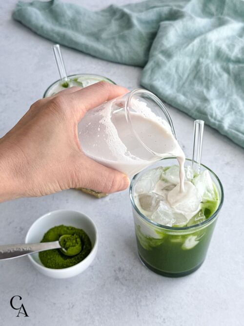 A woman's hand pouring almond milk over a cup of matcha with ice.