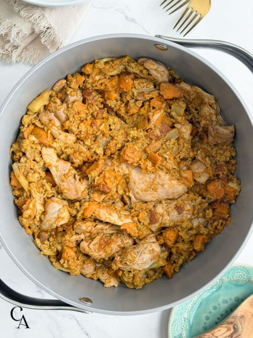 A pot of vindaloo chicken thighs with sweet potato and cauliflower, next to a turquoise plate with a wooden spoon.