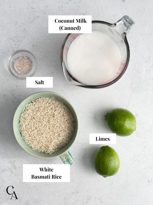 A measuring cup of coconut milk, a bowl of uncooked basmati rice, salt and two limes on a light gray countertop.