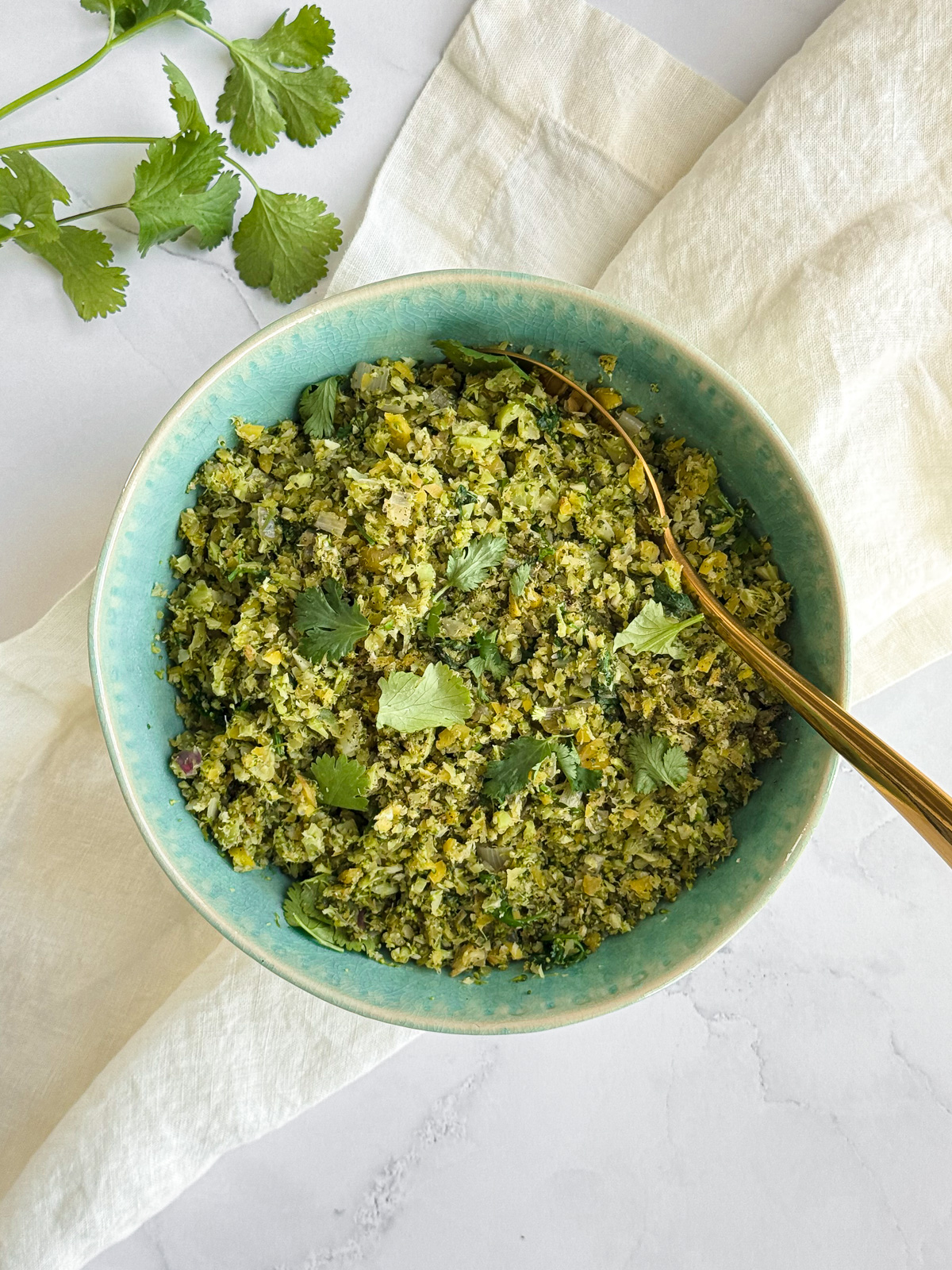 A bowl of broccoli rice on a linen napkin with a gold serving spoon and sprigs of fresh cilantro.
