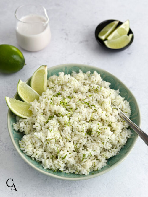A bowl of coconut and lime flavored basmati rice with a pitcher of coconut milk and fresh limes in the background.