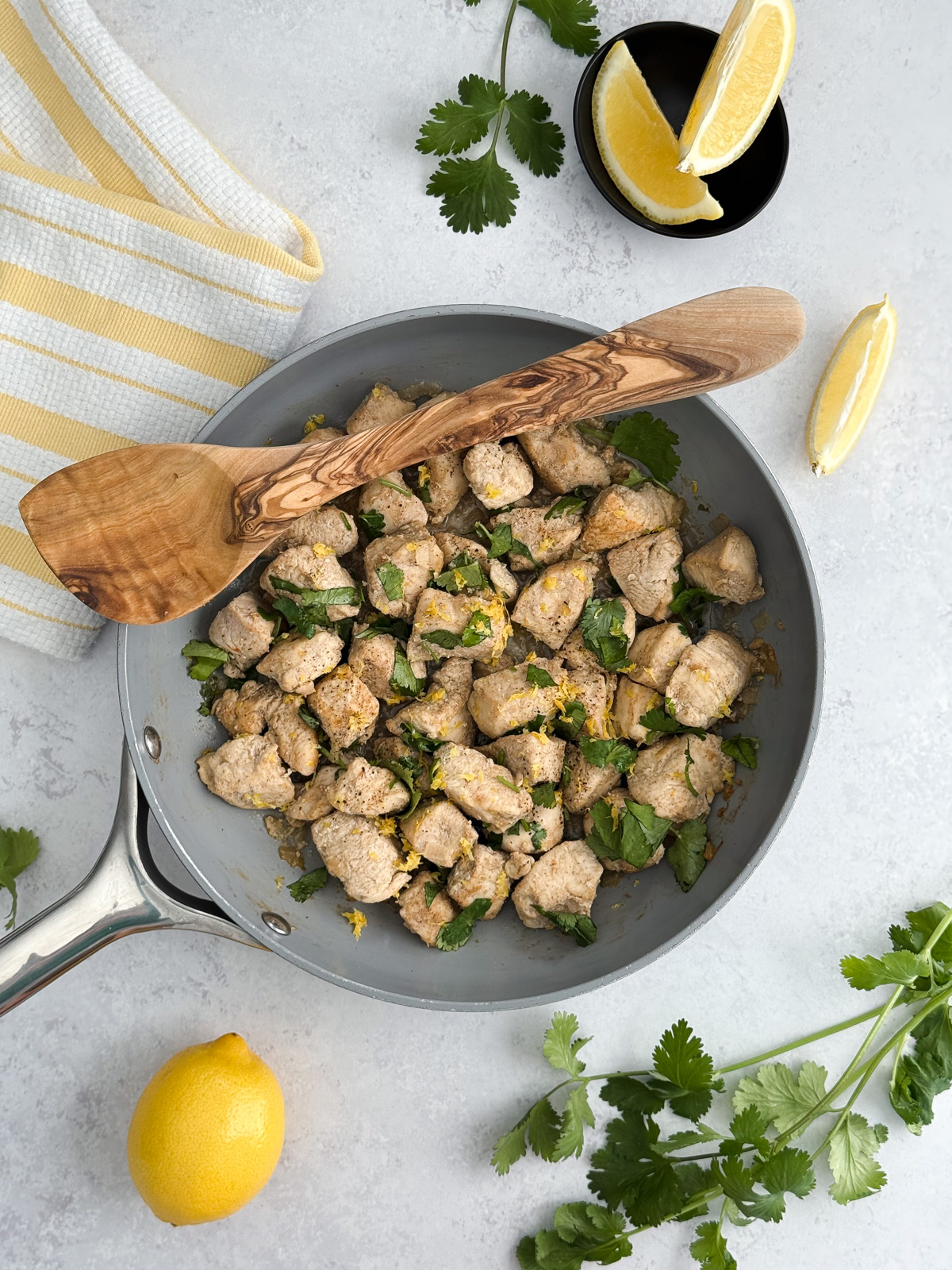 A pan of cooked chicken breasts with a bunch of cilantro and a bowl of lemon wedges sitting on a light counter.