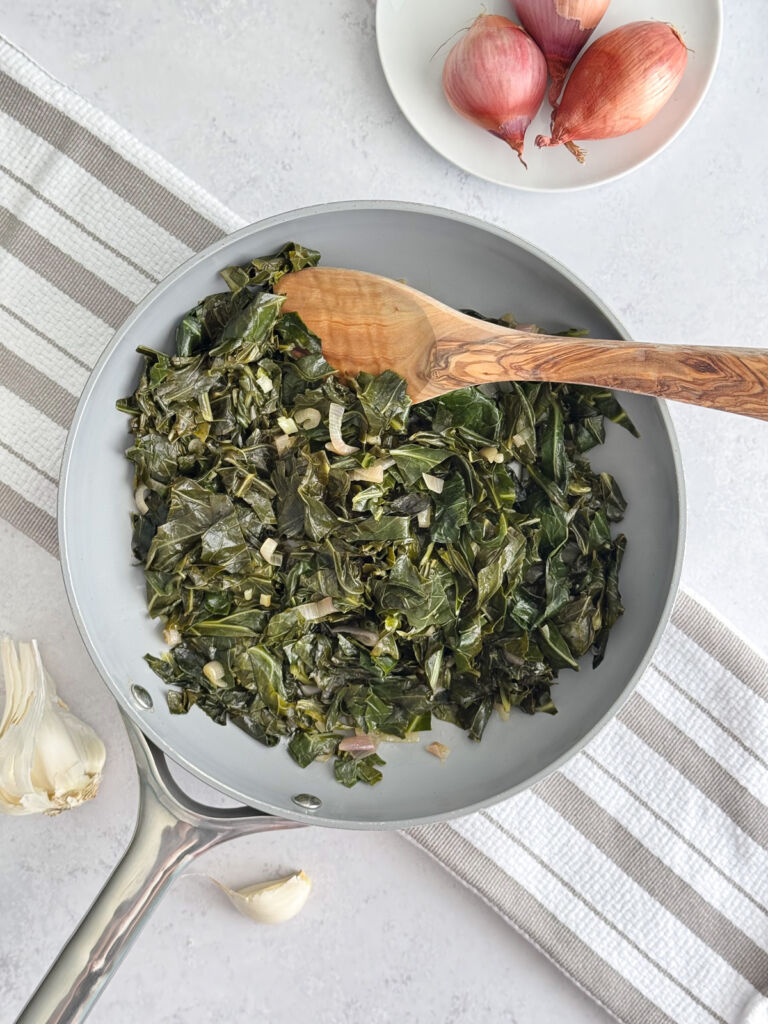 Sautéed collards in a pan with a wooden spoon on top of a striped towel.