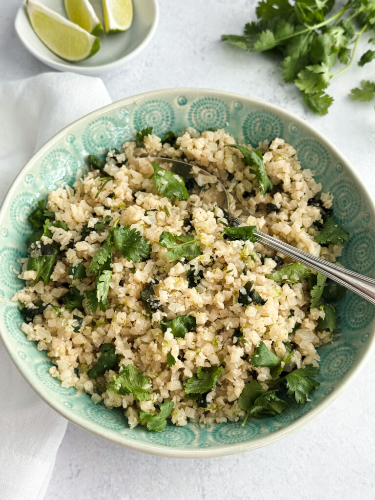A bowl of cauliflower rice with fresh cilantro, lime wedges, and lime zest.