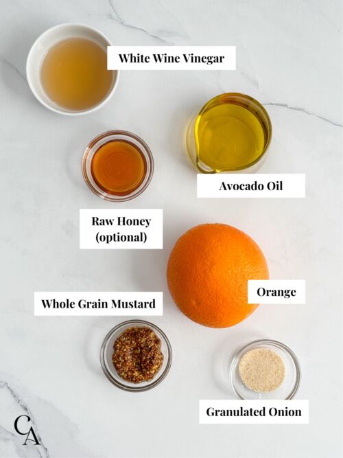 Ingredients on a marble countertop, including an orange, mustard, avocado oil, and vinegar.