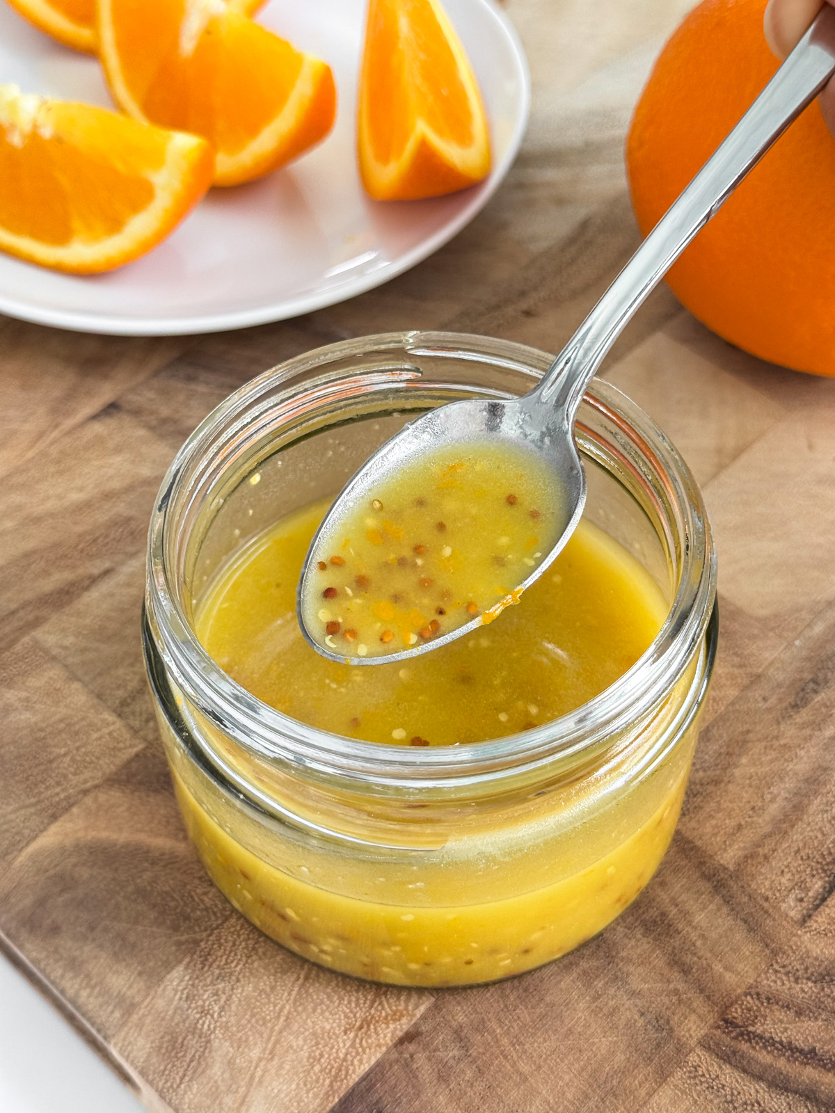 A hand spooning a jar of orange vinaigrette sitting on a wooden cutting board.