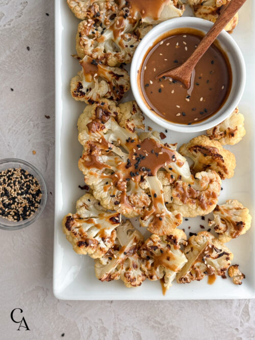 Roasted cauliflower steaks on a white platter, drizzled with maple tahini dressing and sprinkled with black and white toasted sesame seeds.