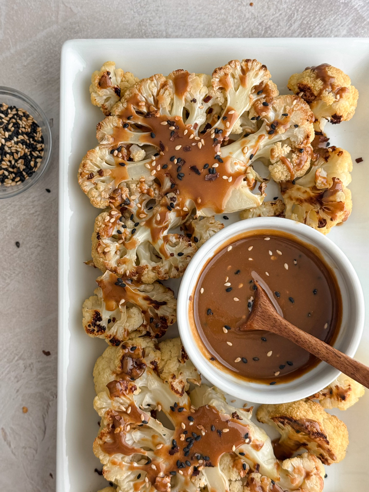 Golden cauliflower steaks topped with maple tahini dressing and sesame seeds, served with extra sauce in a white bowl and a wooden spoon.