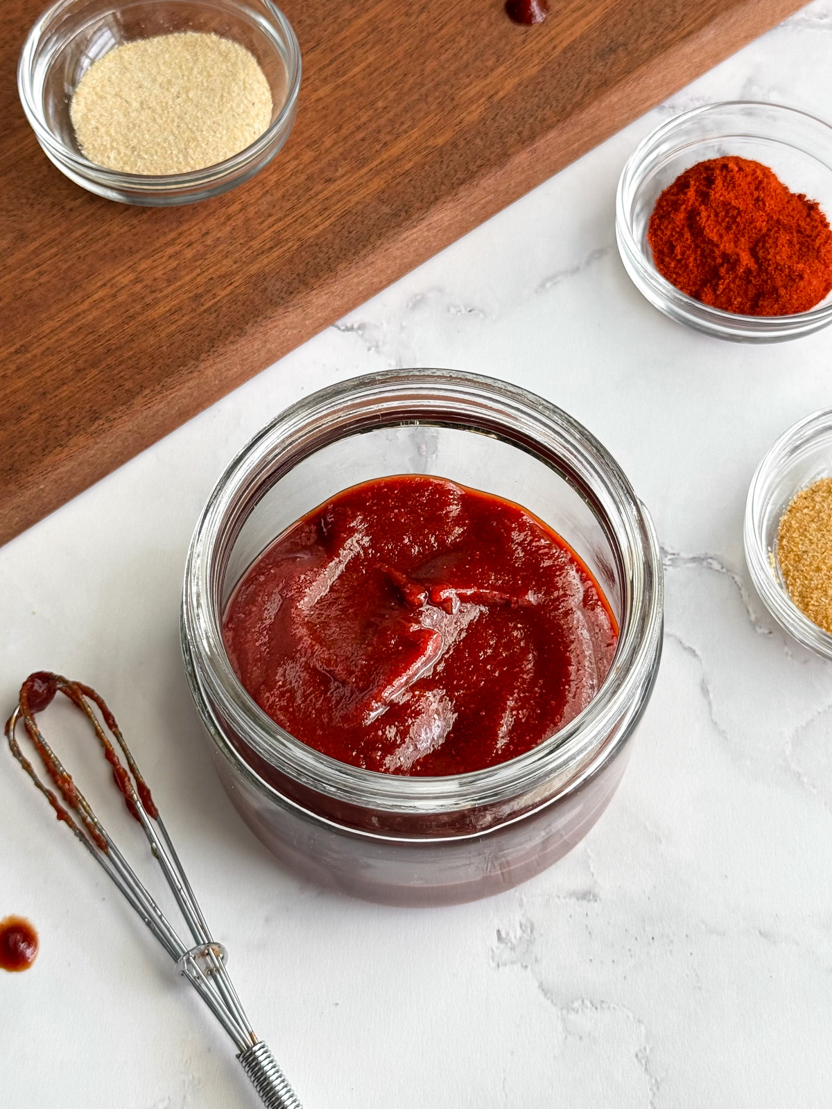 A container of homemade honey barbecue sauce with granulated onion, paprika, and garlic on a stone top.