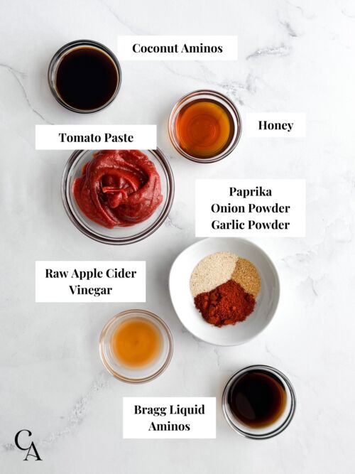 Bowls of spices, aminos, honey, and tomato paste on a white surface.