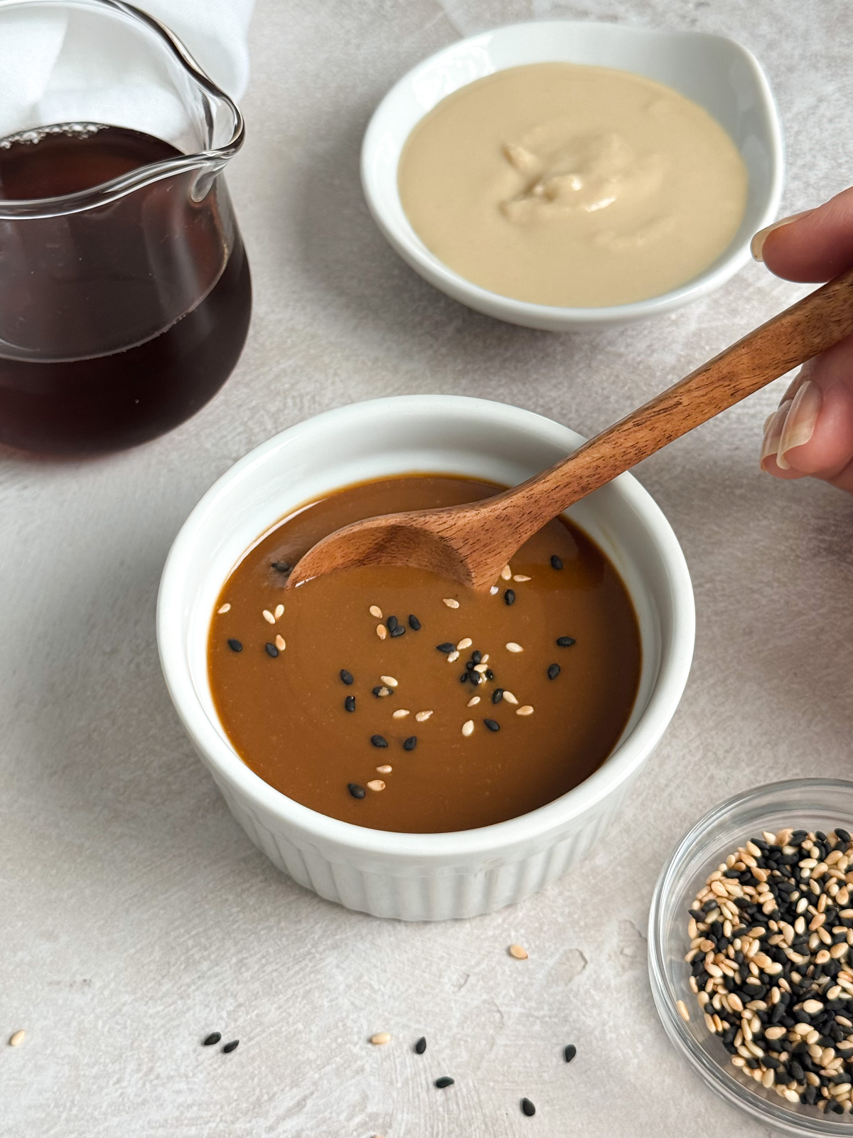 A bowl of maple tahini dressing with sesame seeds, a bowl of tahini paste, and a woman's hand holding a spoon.
