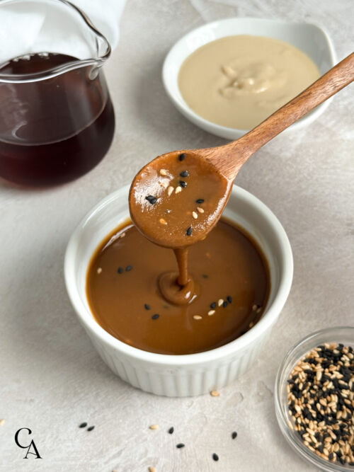 A spoonful of dark tahini sauce drizzling over a ramekin, with a bowl of sesame seeds.