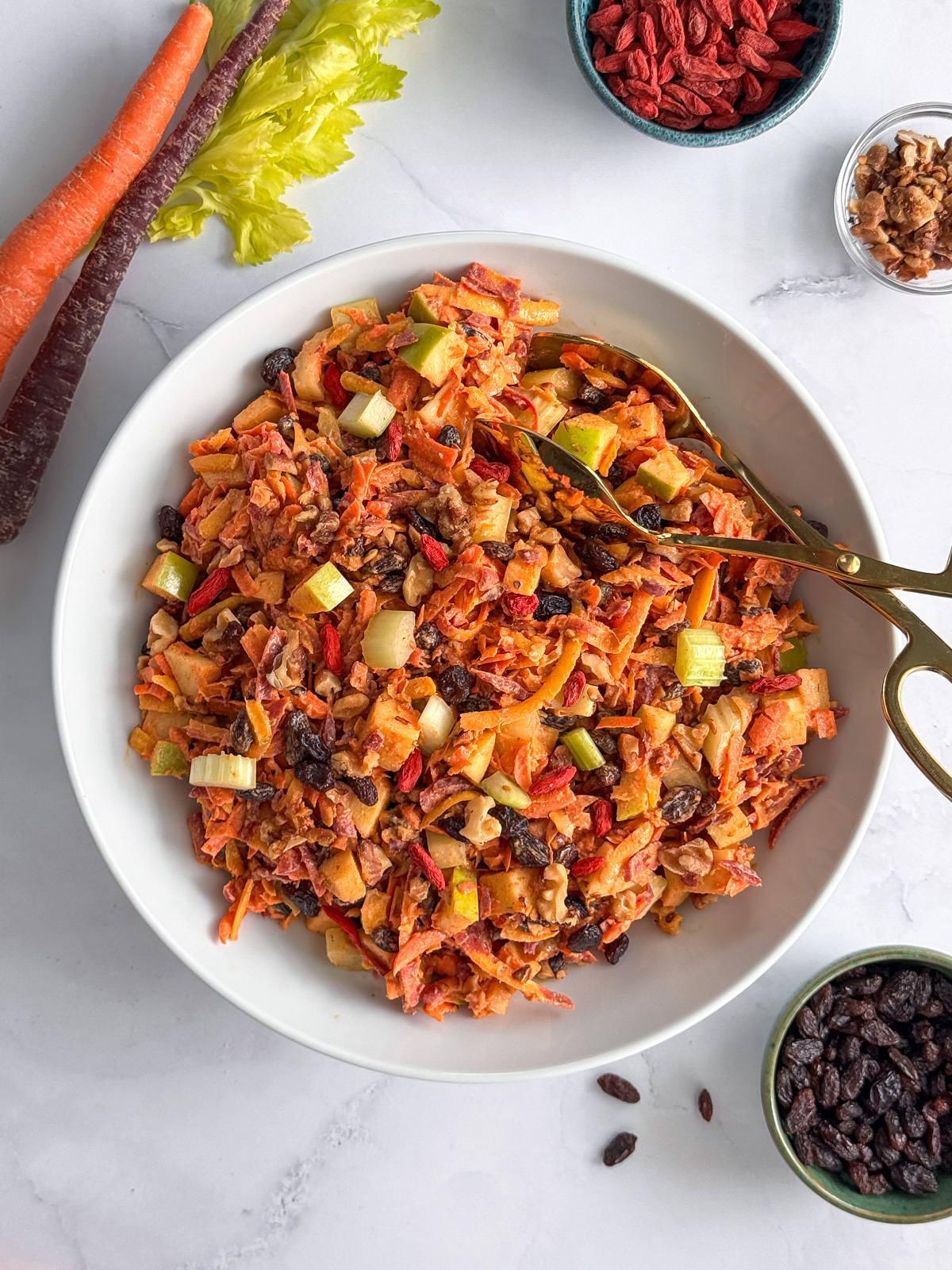 Overhead shot of a fruit salad with shredded rainbow carrots, raisins, celery, and apples with gold tongs and bowls of nuts on the side.