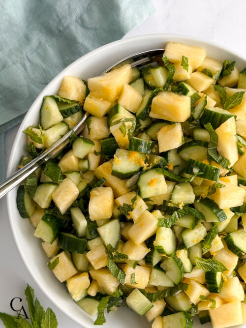 Chopped pineapple and english cucumber slices with chopped mint leaves and lemon zest in a white serving bowl.