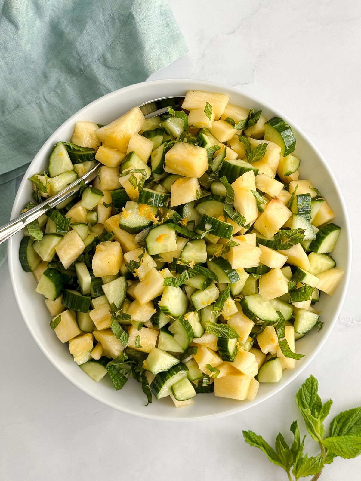 A large white bowl with chopped pineapple, English cucumber and fresh mint leaves.