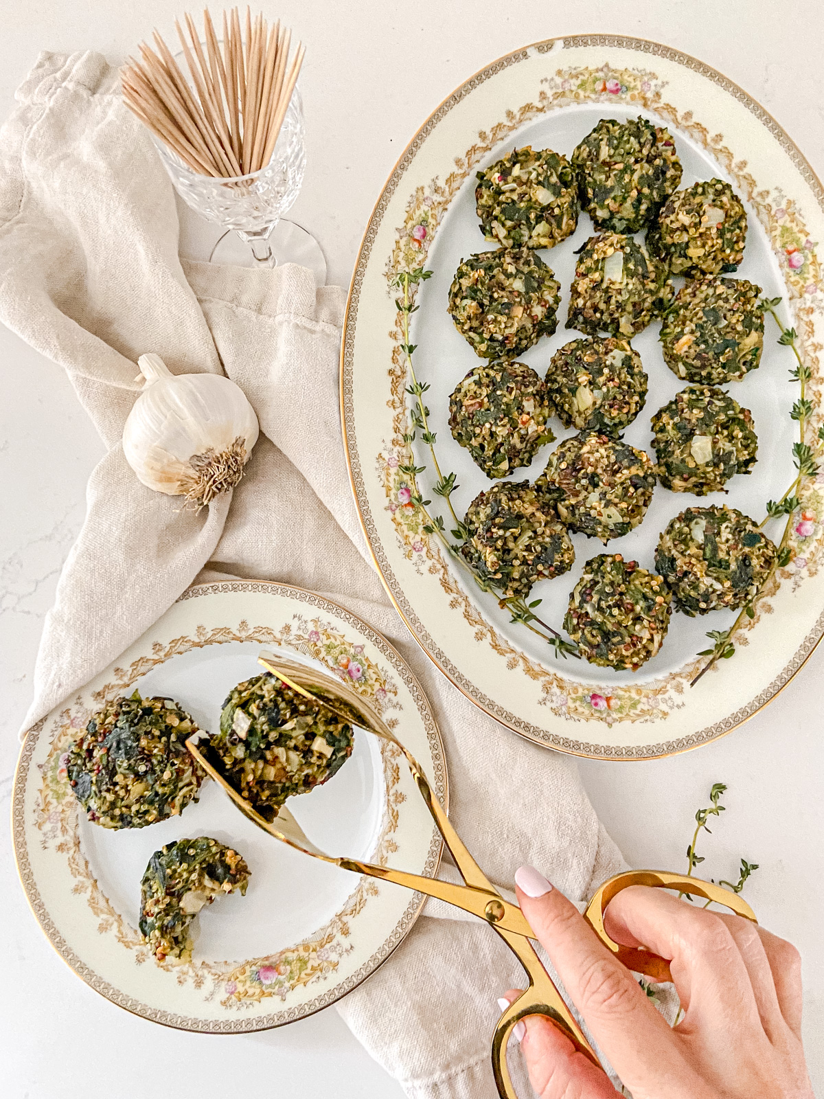 A woman's hand holding gold tongs over a platter of spinach balls with a cup of toothpicks nearby.
