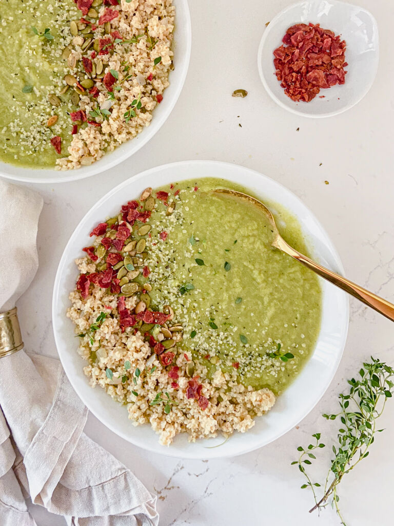 White bowls of creamy broccoli soup with fresh herbs, pepitas, and cooked millet on top, with a bowl of bacon bits nearby.