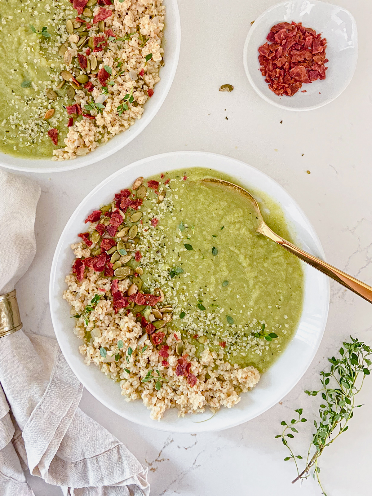 White bowls of creamy broccoli soup with fresh herbs, pepitas, and cooked millet on top, with a bowl of bacon bits nearby.