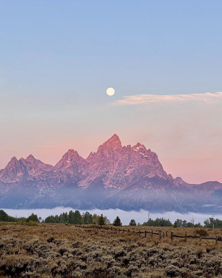 A landscape outside of Jackson Hole, with the full moon over the Tetons behind a field with a wooden fence.