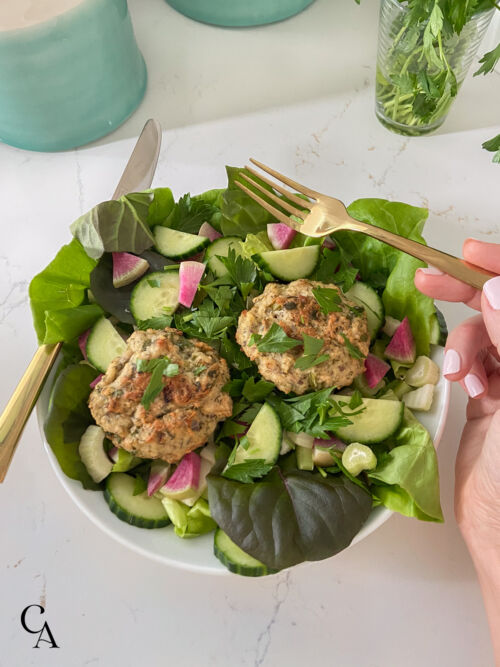 A green salad with sliced cucumbers, watermelon radishes, and salmon patties with fresh parsley sprinkled on top.