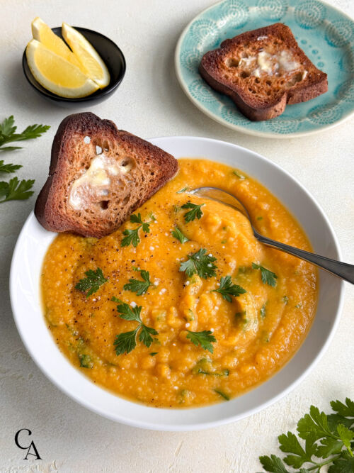 Bowl of roasted butternut squash soup topped with parsley, served with lemon wedges and a slice of buttery toast on a turquoise plate.
