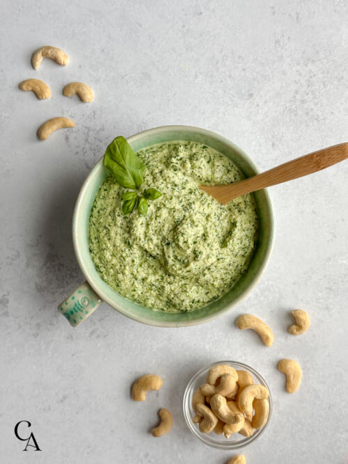 A turquoise bowl of creamy pesto with cashews next to it and a bamboo serving spoon.