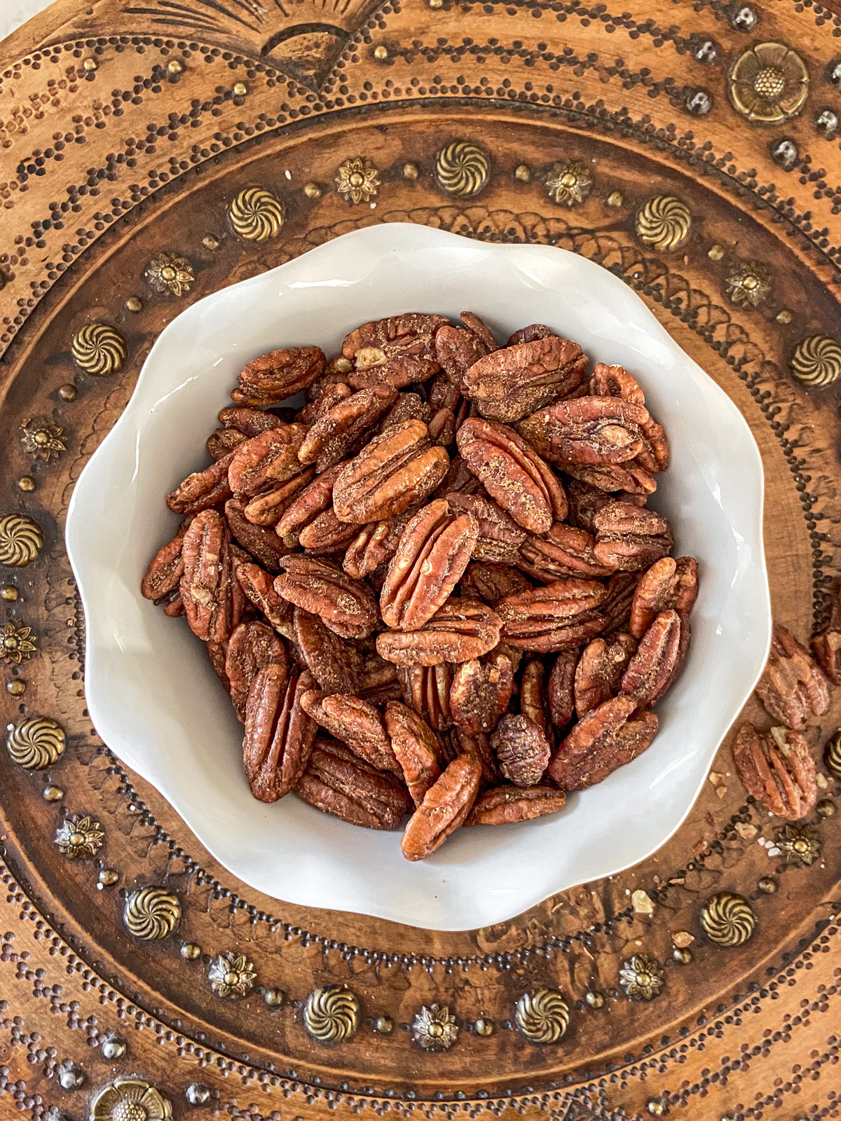 A small, white bowl of candied nuts on an antique breadboard.