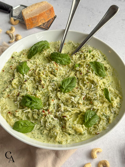 Close-up of a bowl of creamy pesto spaghetti squash, garnished with fresh basil and shredded cheese, next to a block of Parmegiano-Reggiano and cashews.