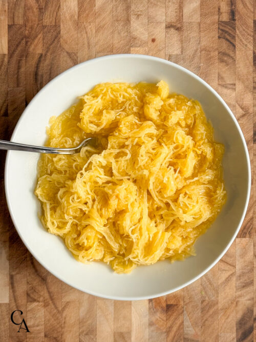 A white bowl of spaghetti squash, on a dark cutting board.