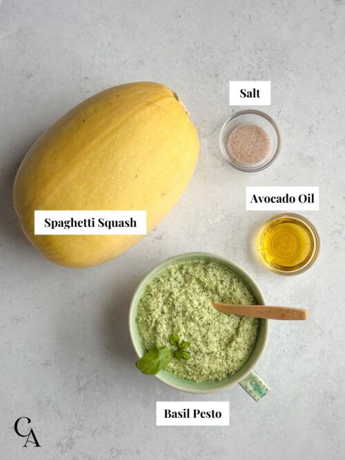 Flat lay of labeled ingredients for pesto spaghetti squash, including a whole squash, salt, avocado oil, and a bowl of creamy basil pesto.