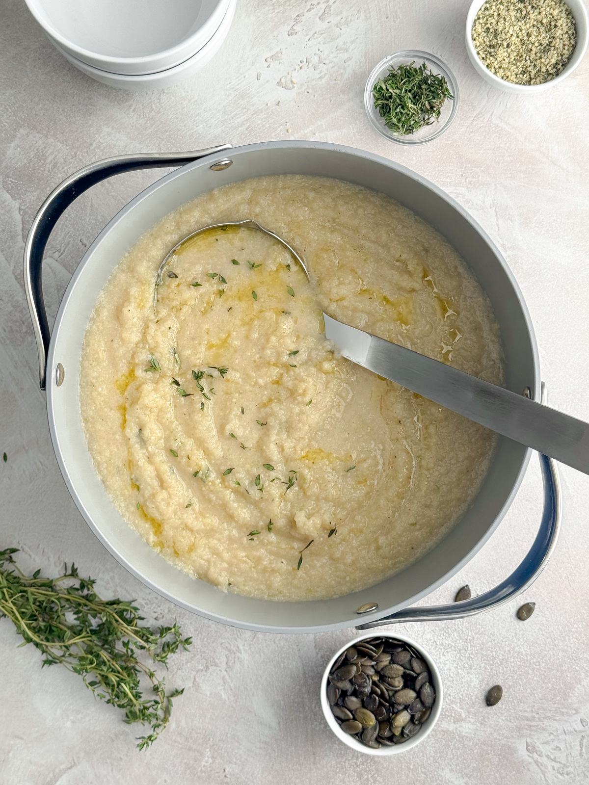 Overhead view of a pot filled with cauliflower soup, with a metal ladle inside and bowls of herbs and seeds around it.