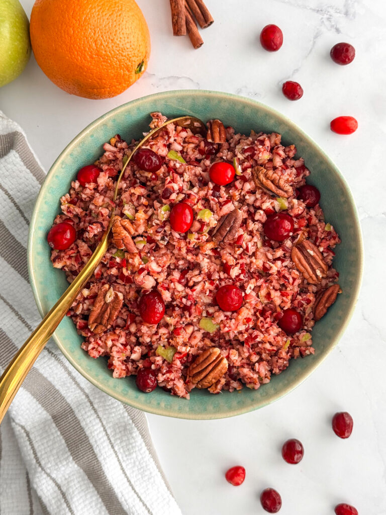 Top-down view of cranberry orange relish in a teal bowl, with scattered cranberries and whole fruit nearby on a marble surface.