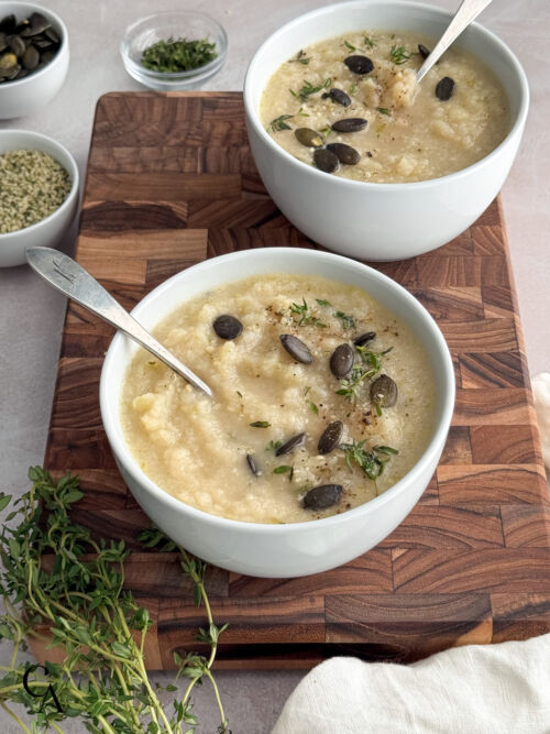 Two white bowls filled with cauliflower soup sit on a wooden board. Each bowl is topped with pumpkin seeds and fresh herbs.