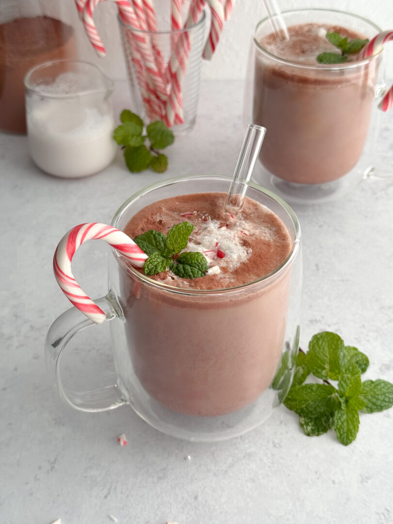 Close-up of a festive glass mug filled with hot cocoa, garnished with crushed candy cane, fresh mint, and a glass straw.