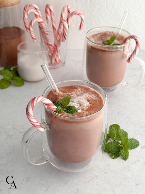 Two glass mugs of peppermint hot cocoa topped with mint, surrounded by fresh mint, a cup of candy canes, and a pitcher of almond milk.