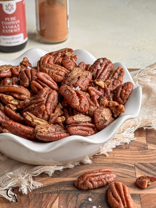 A small pie dish full of candied pecans, sprinkled with flaky salt, with jars of spices in the background.