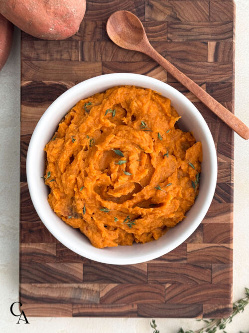 Overhead view of sweet potato mash in a white bowl on a wooden board, garnished with herbs and served with a wooden spoon.