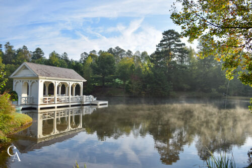 A white boathouse on a pond with mist rising off the water at sunrise.