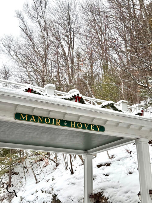 A painted wooden sign that says Manoir Hovey, hanging from a snow-covered porte cochere.