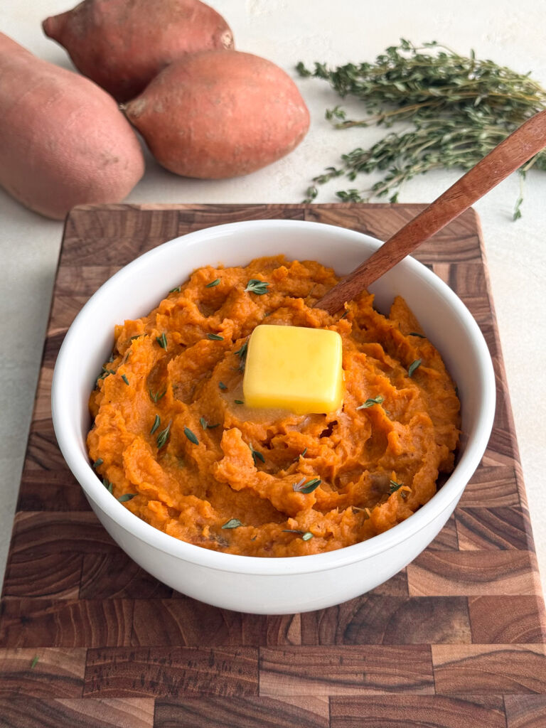 Creamy mashed sweet potatoes topped with a pat of butter, served in a white bowl on a wooden board, with fresh thyme and whole sweet potatoes in the background.