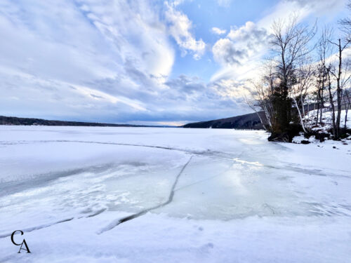 A frozen Canadian lake with bare trees along the shore and blue skies above.